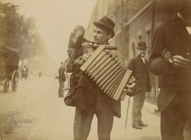 A man playing an accordion with a monkey sitting on his shoulder. The man is wearing a hat and is looking towards the monkey. The monkey has a paw over its face.