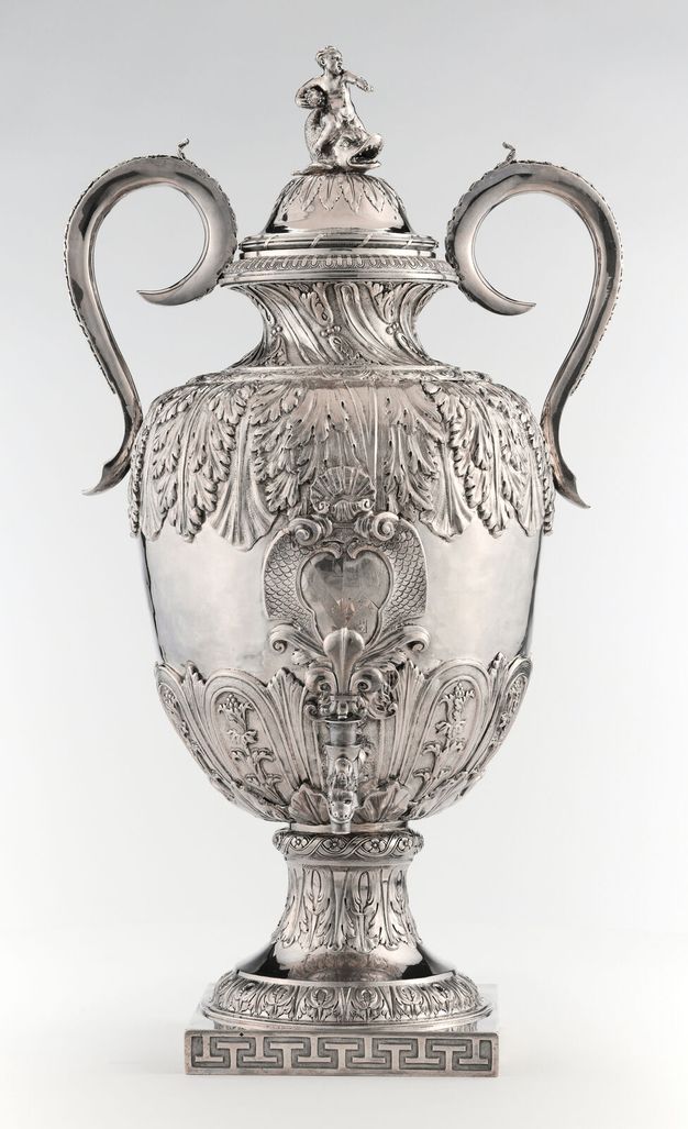 Symmetrical silver urn with two large handles on either side at the top, a water spigot on the front, and the face of a lion in relief on the back.