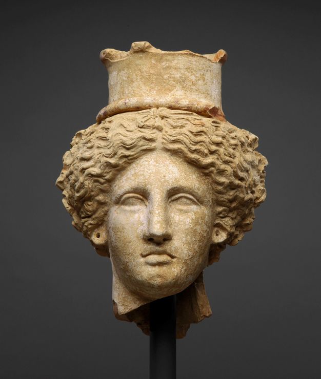 Clay bust of a female head wearing a polos, which is decorated on the upper edge with small protruding nodes. The figure’s thick wavy hair is divided symmetrically into two masses and pulled back.
